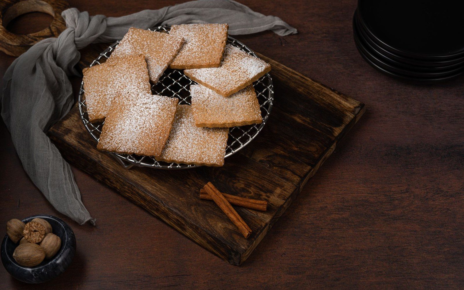 Square biscochitos dusted with powdered sugar on a wire rack beside cinnamon sticks