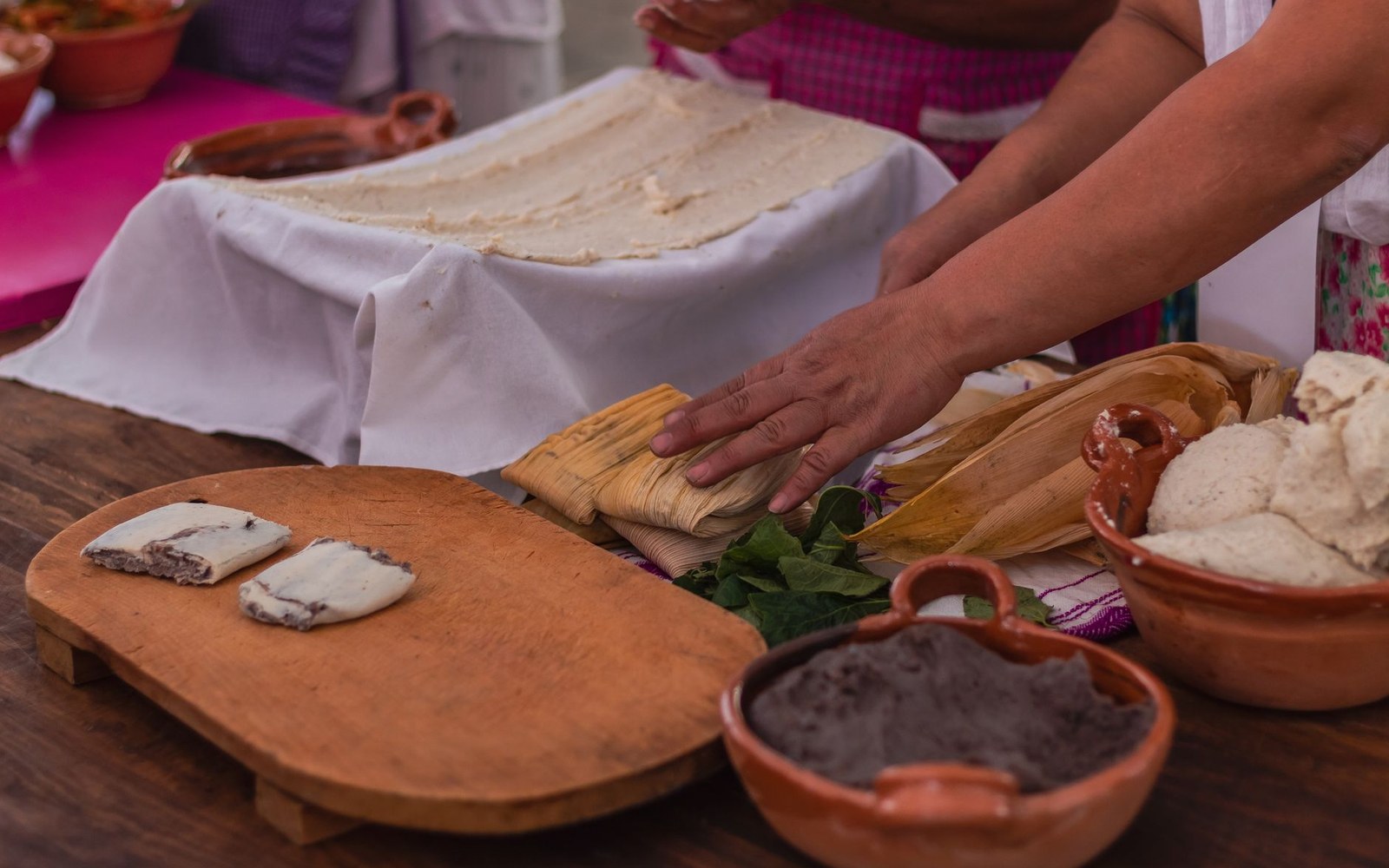 Hands wrapping tamales in corn husks beside bowls of masa and mole filling