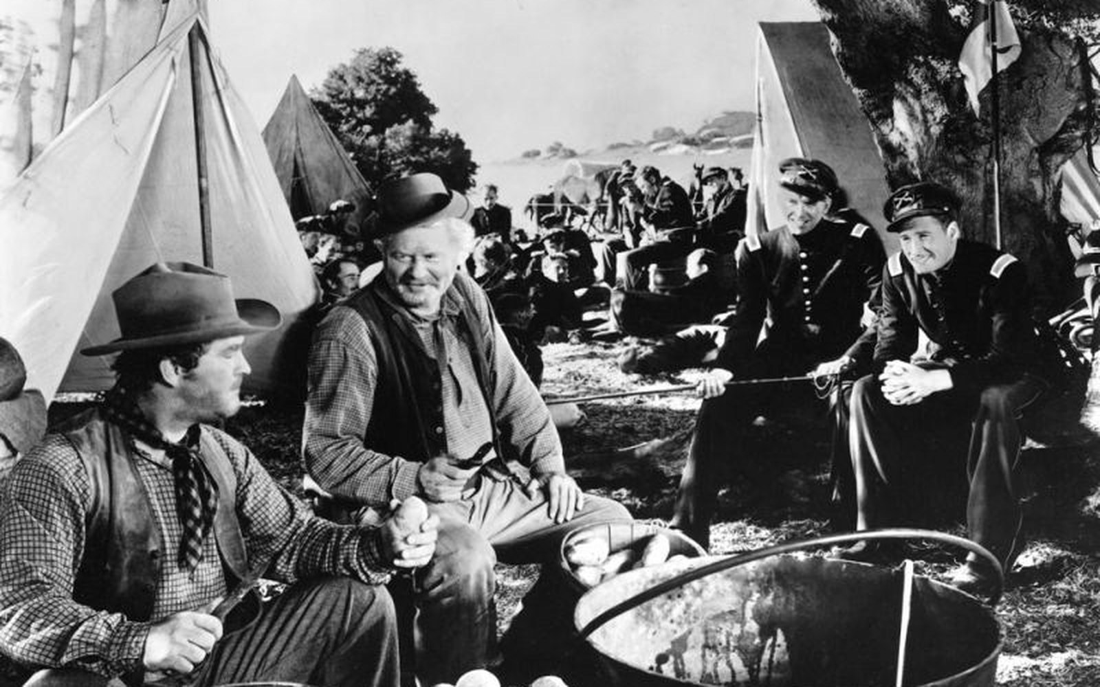 Men in cavalry uniforms and civilian clothes sit around a cooking pot near tents